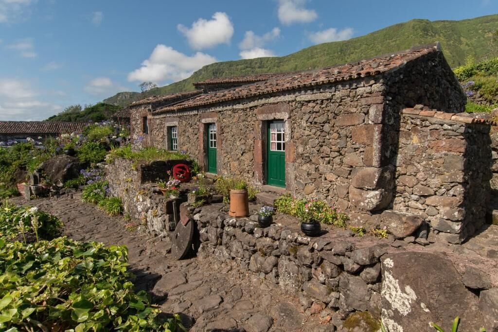 Un bâtiment en pierre, doté d’un toit en tuiles, entouré de verdure et agrémenté de plantes en pot.