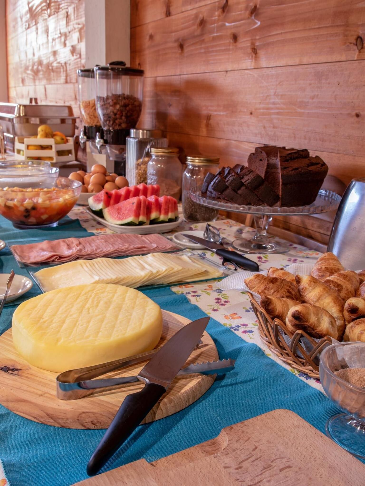 A table with various food items including a large wheel of cheese, sliced bread, pastries, and a watermelon.