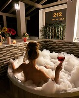 Una persona relajándose en un jacuzzi con una copa de vino tinto, rodeada de flores y un cartel que indica el lugar como Hotel Spa 212.