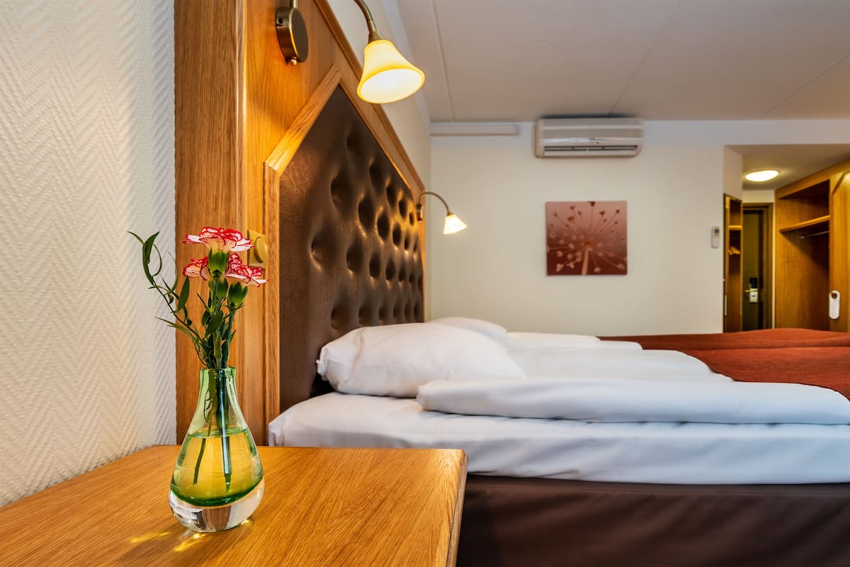 A hotel room with a wooden headboard, a bedside table with a vase of flowers, neatly made beds, and a wall-mounted air conditioner.