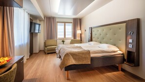 Habitación de hotel con cama, una silla, televisor, ventana con cortinas y una lámpara.
