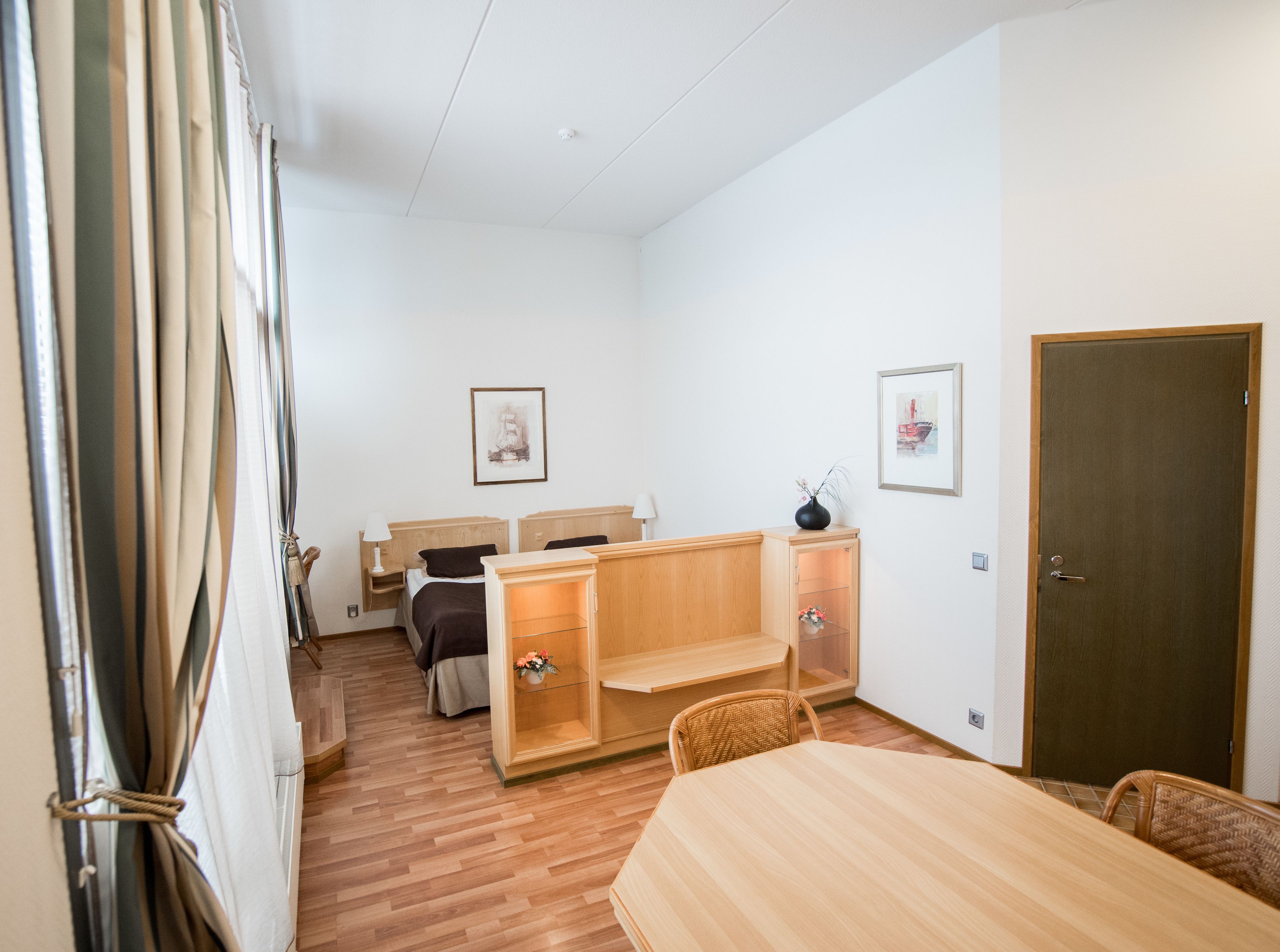 A room with a bed, bedside table, lamp, wooden cabinet, and a dining table with chairs.