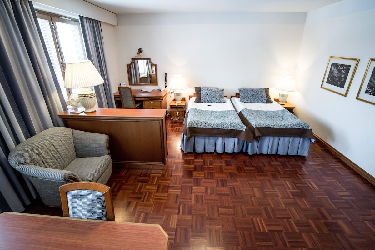 A hotel room with two beds, a desk, a chair, and a lamp.