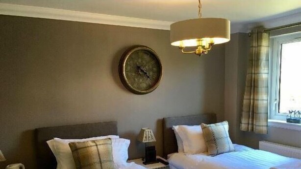 A hotel room with two beds, a clock on the wall, a lamp, and a window with curtains.