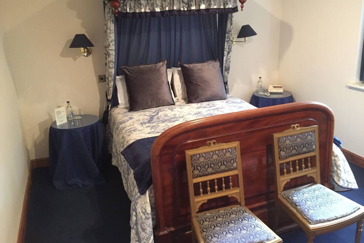 A four-poster canopy bed with a floral bedspread, two bedside tables with lamps, and a small wooden chair with a patterned cushion.