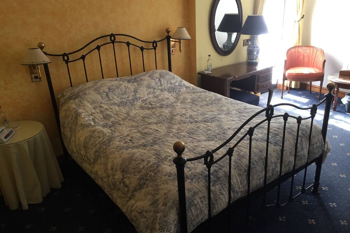 A bed with a metal frame and a floral bedspread, a bedside table with a lamp, a chair, and a mirror on the wall.