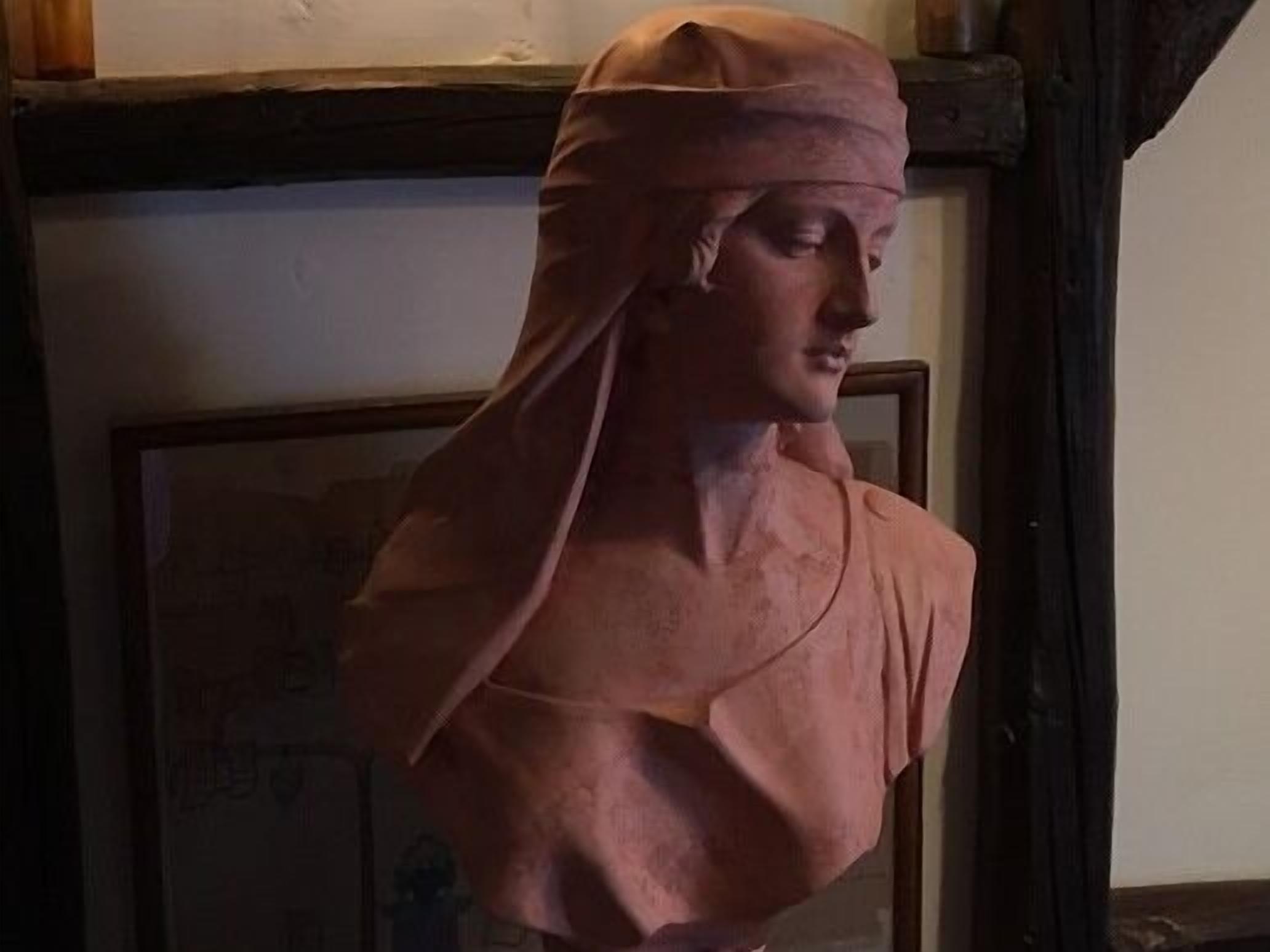 Une sculpture en buste se trouve dans une pièce où est accrochée une carte encadrée au mur, et une poutre en bois surplombe l’ensemble.