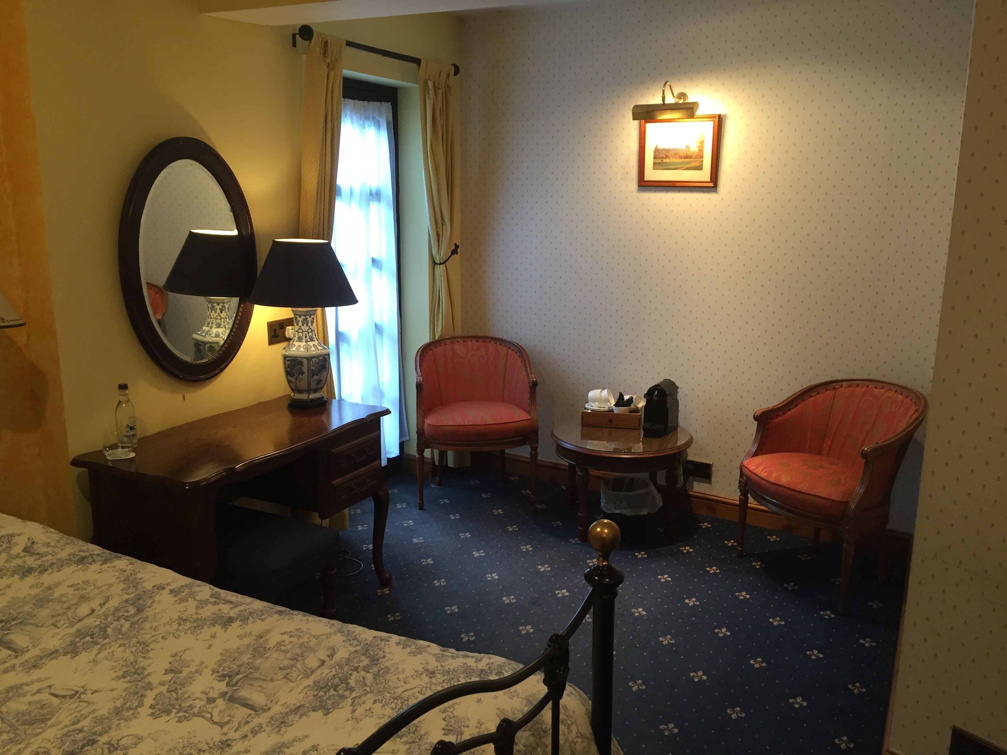 A hotel room with a bed, two armchairs, a small table with a lamp, a mirror, and a picture on the wall.