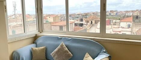 A room with a blue sofa, two pillows, and a large window with a city view.