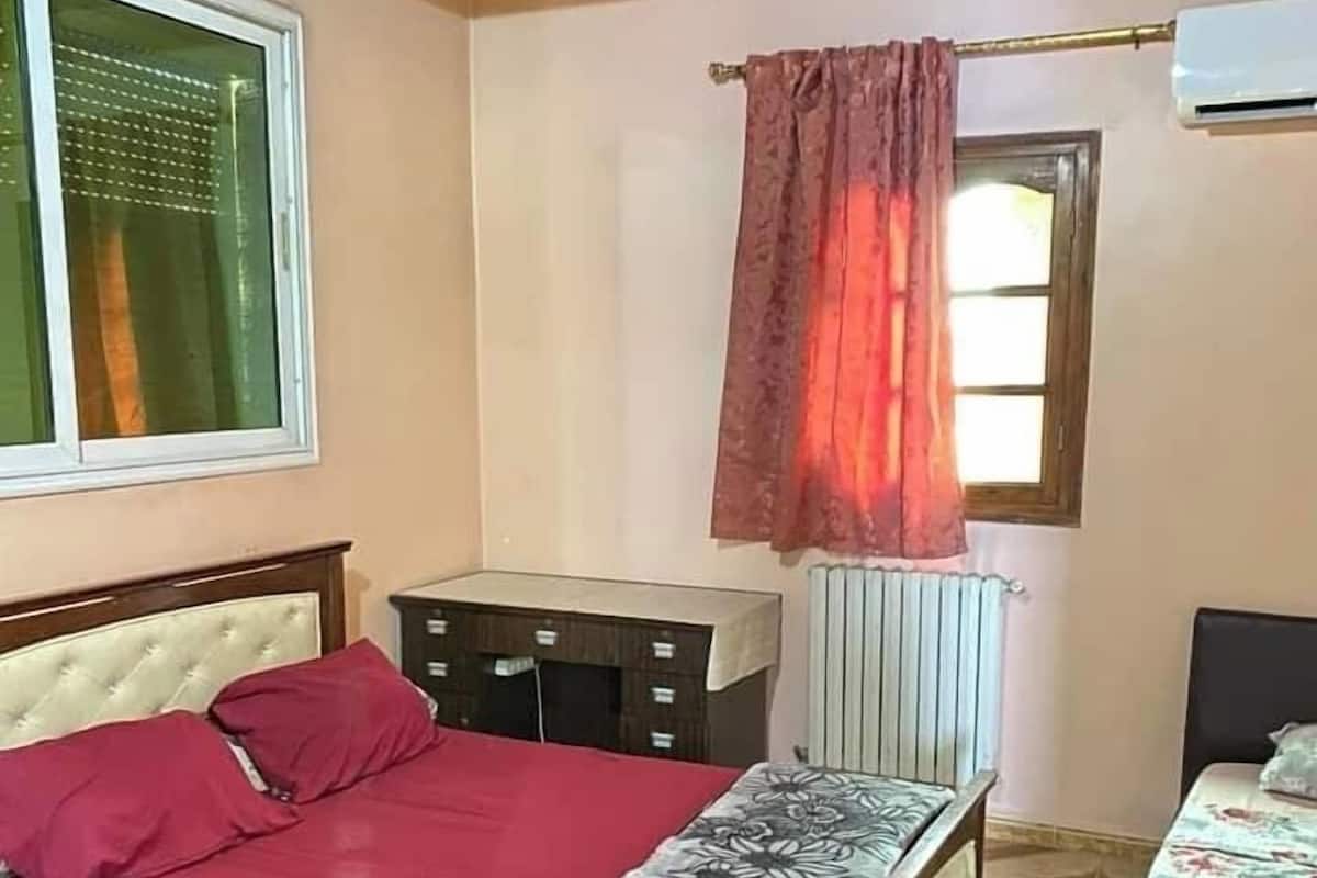 A bedroom with a bed, a nightstand, a radiator, a window with curtains, and a wooden ceiling.