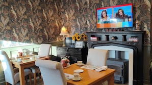 A dining area with a wooden table and chairs, a television displaying a news program, a fireplace, and a patterned wallpaper. - The Corner House (Lowestoft)