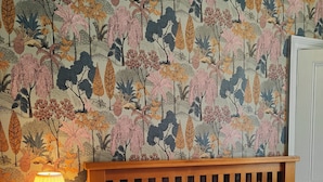 A bedroom with a wooden headboard, a bedside table with a lamp, and a floral wallpaper. - The Corner House (Lowestoft)