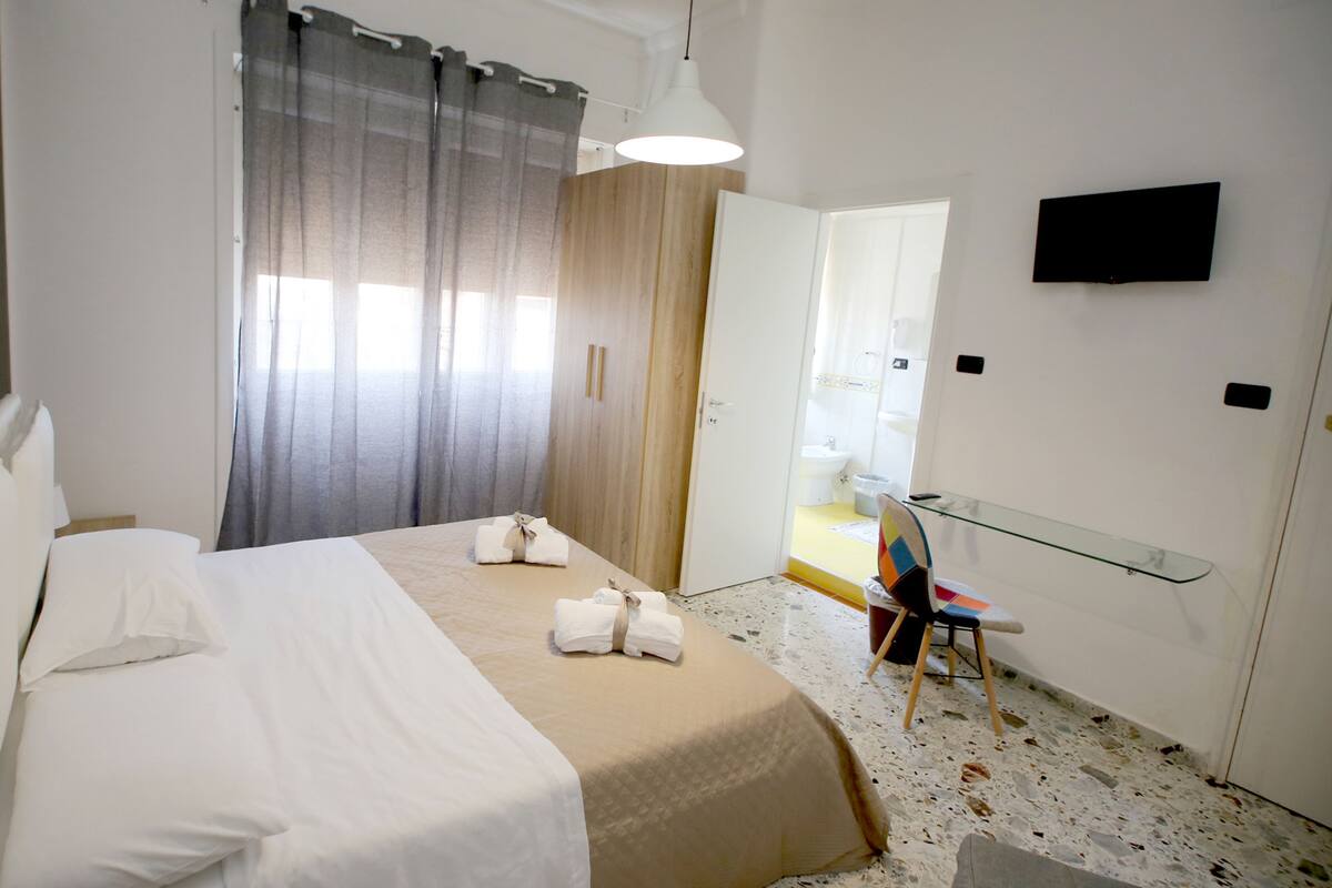 A hotel room with a bed, a glass side table, a chair, and a television on the wall.
