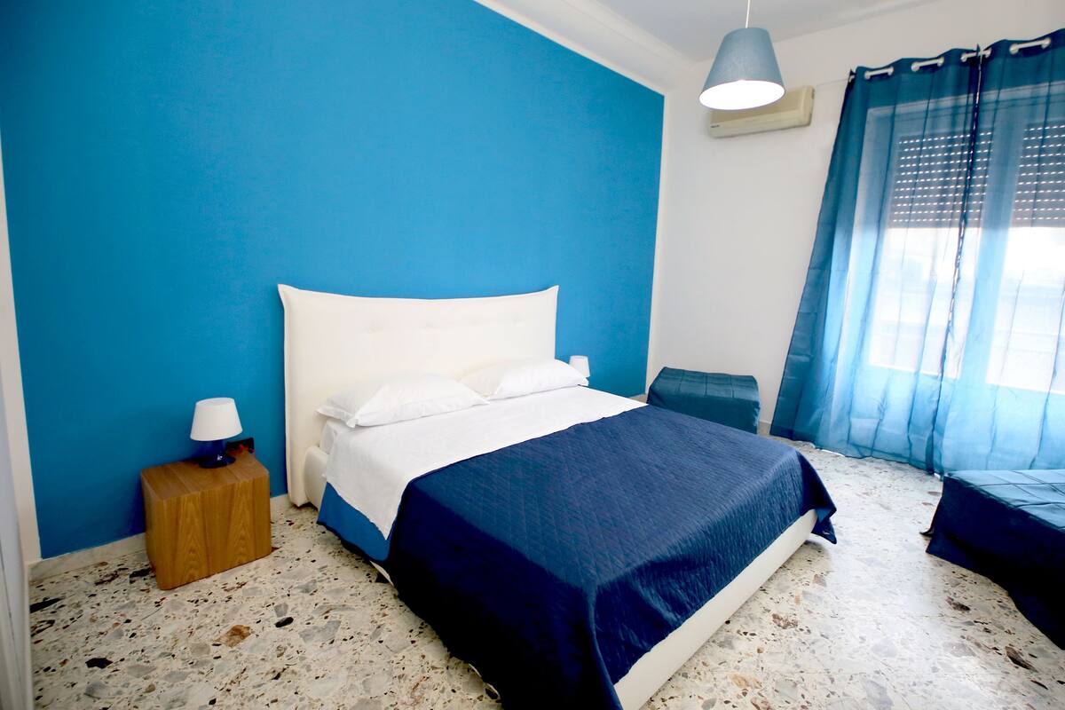 A bedroom with a bed, a nightstand, a lamp, a window with curtains, and a wall painted blue.