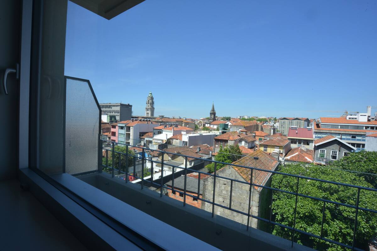 A view from a balcony with a metal railing, overlooking a cityscape with various buildings and a prominent tower.