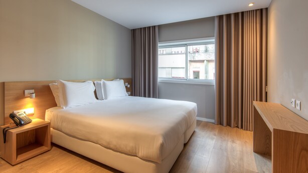 A hotel room with a large bed, bedside table with a phone, a small desk, and a window with curtains.