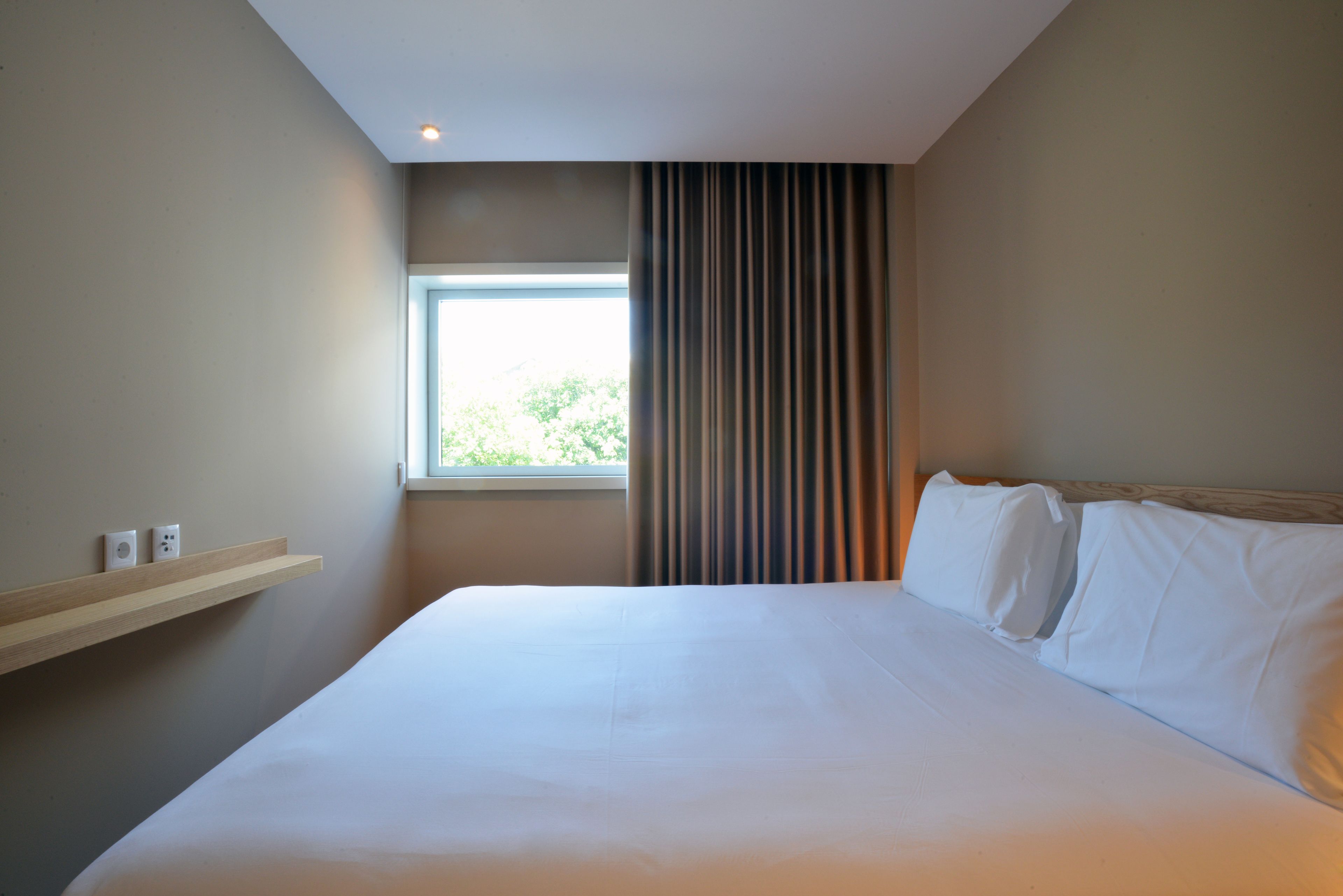 Une chambre d’hôtel avec un grand lit, une fenêtre, une étagère fixée au mur et une prise de courant.