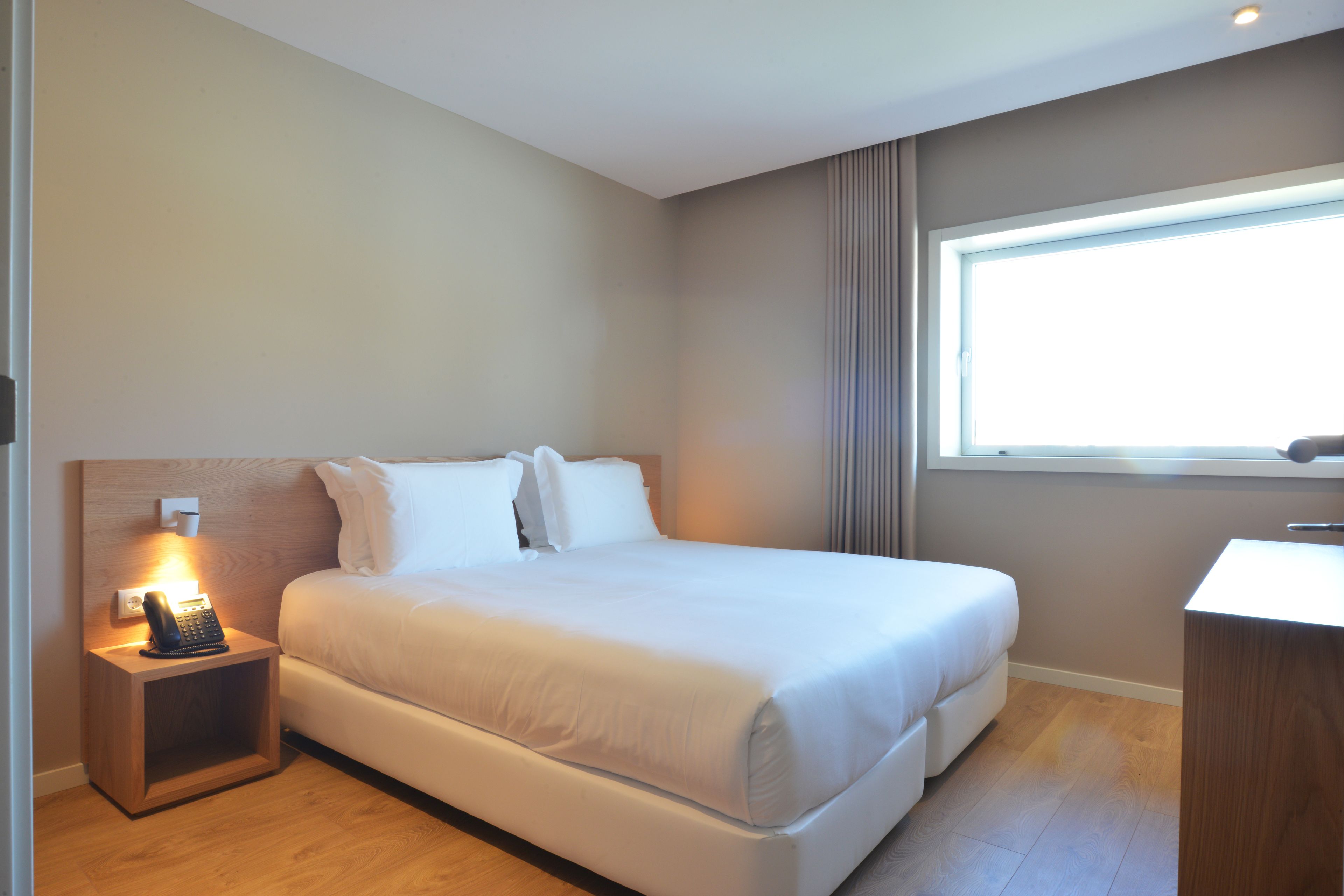 A neatly made bed in a hotel room with a bedside table, a window with curtains, and a wooden nightstand with a phone.