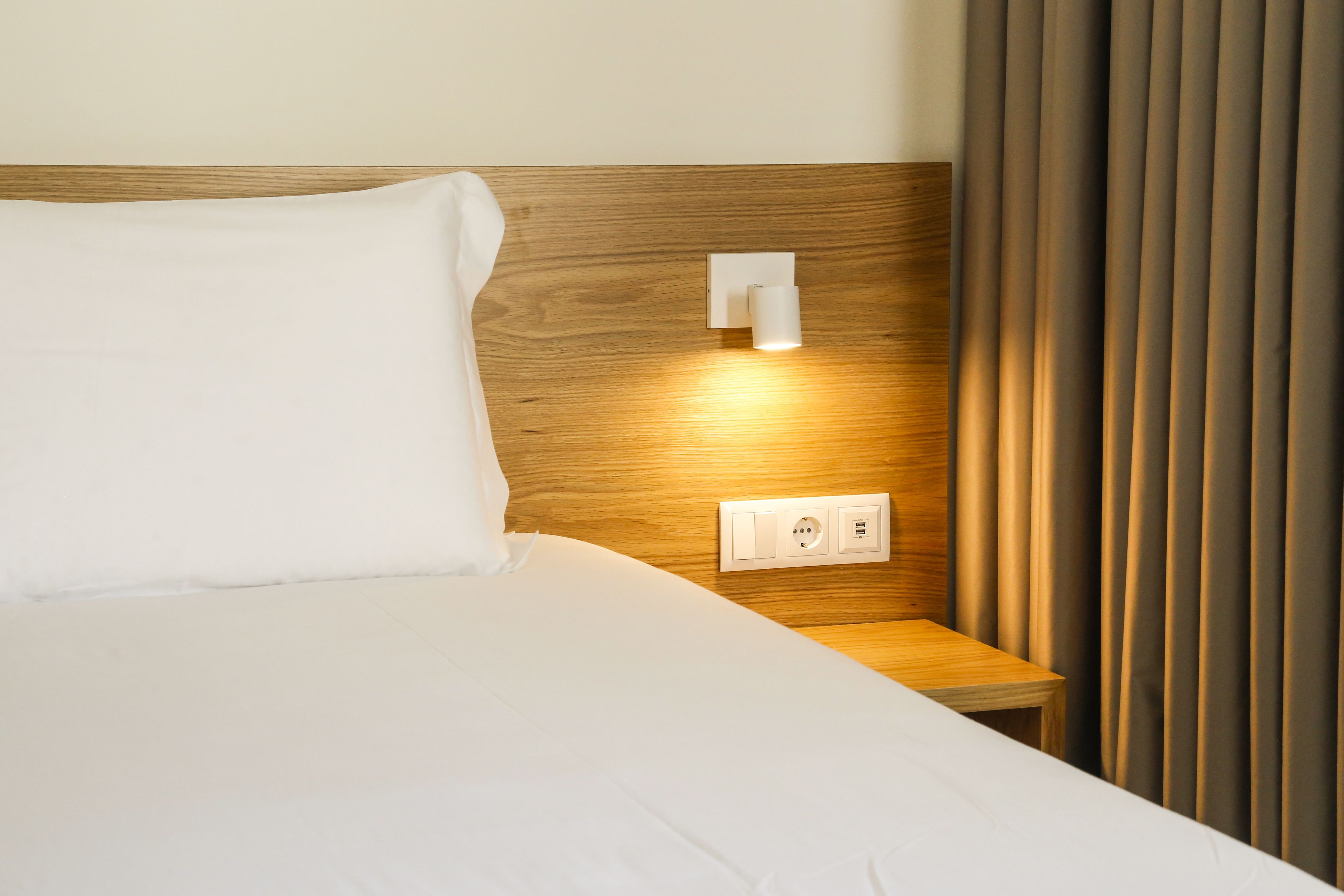 A neatly made bed with white linens, a wooden headboard, a bedside table, and a wall-mounted light fixture.