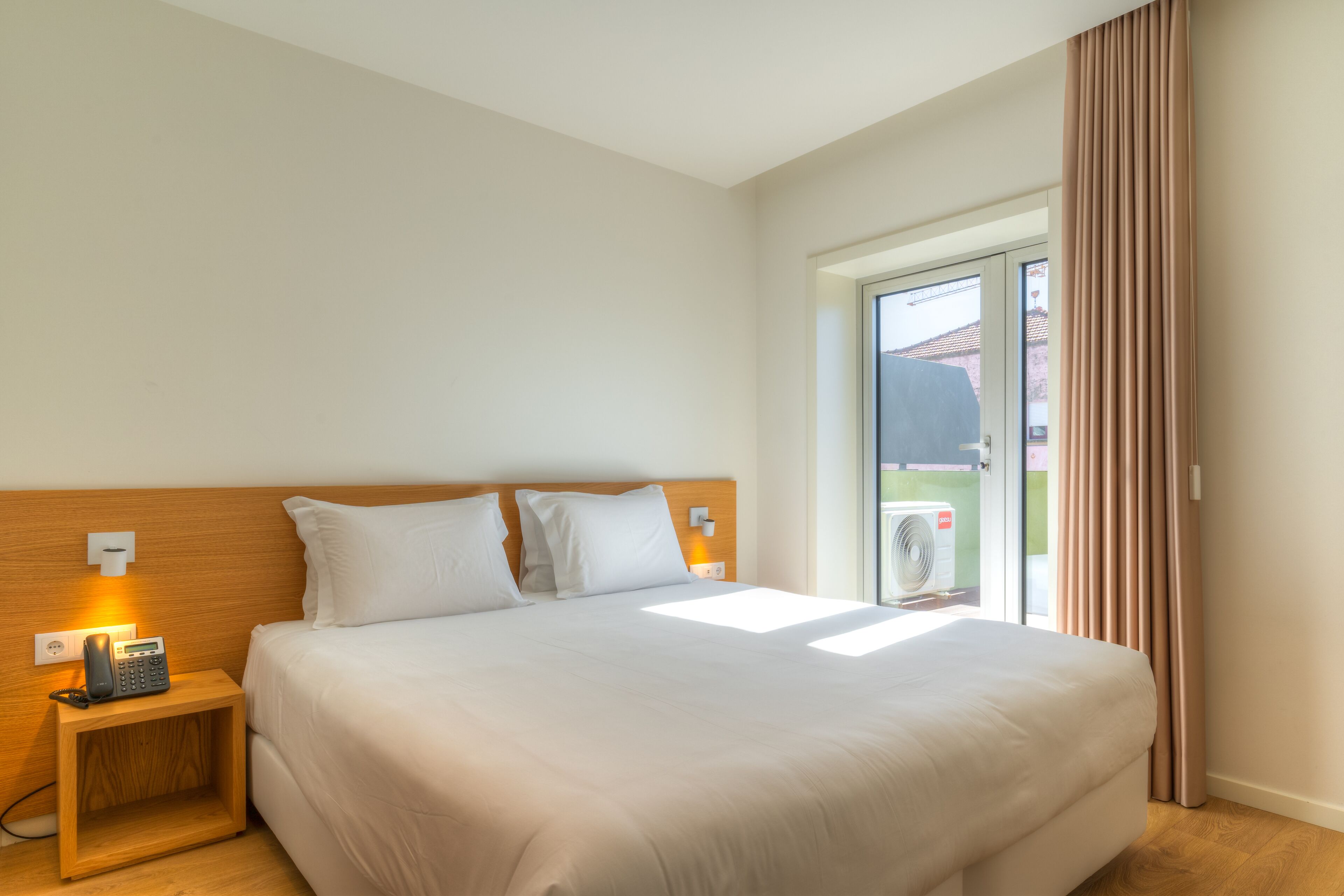 Una habitación de hotel con una cama grande, una mesita de noche con teléfono, una lámpara de pared y una ventana con cortinas.