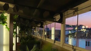 A balcony with a cushioned seating area, potted plants, and a view of the cityscape at dusk. - Usak Yucel Hotel (Usak)