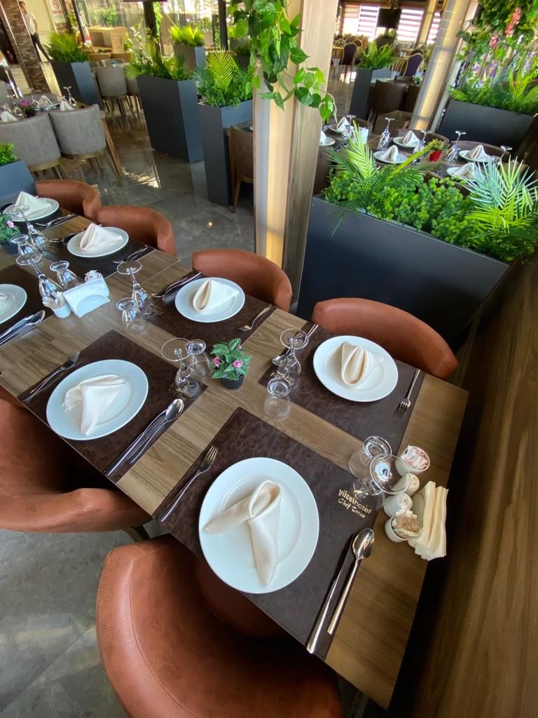 A restaurant setting with a long dining table, chairs, and a central column with greenery.