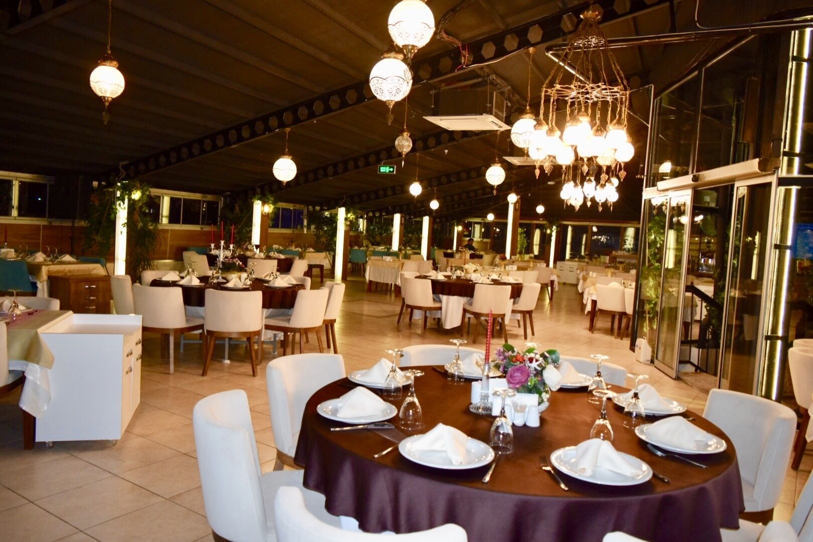 A restaurant with round tables set for dining, featuring white chairs and a dark tablecloth.