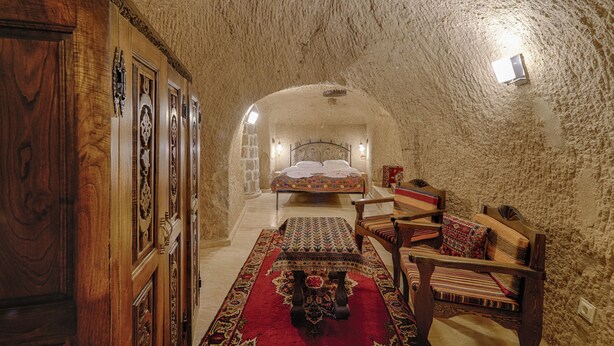 A narrow, stone-walled hallway leading to a bed with a patterned bedspread. A wooden chair and a small table with a red and black patterned tablecloth are visible. - Yoruk Stone House (Avanos)