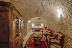 A narrow, stone-walled hallway leading to a bed with a patterned bedspread. A wooden chair and a small table with a red and black patterned tablecloth are visible.