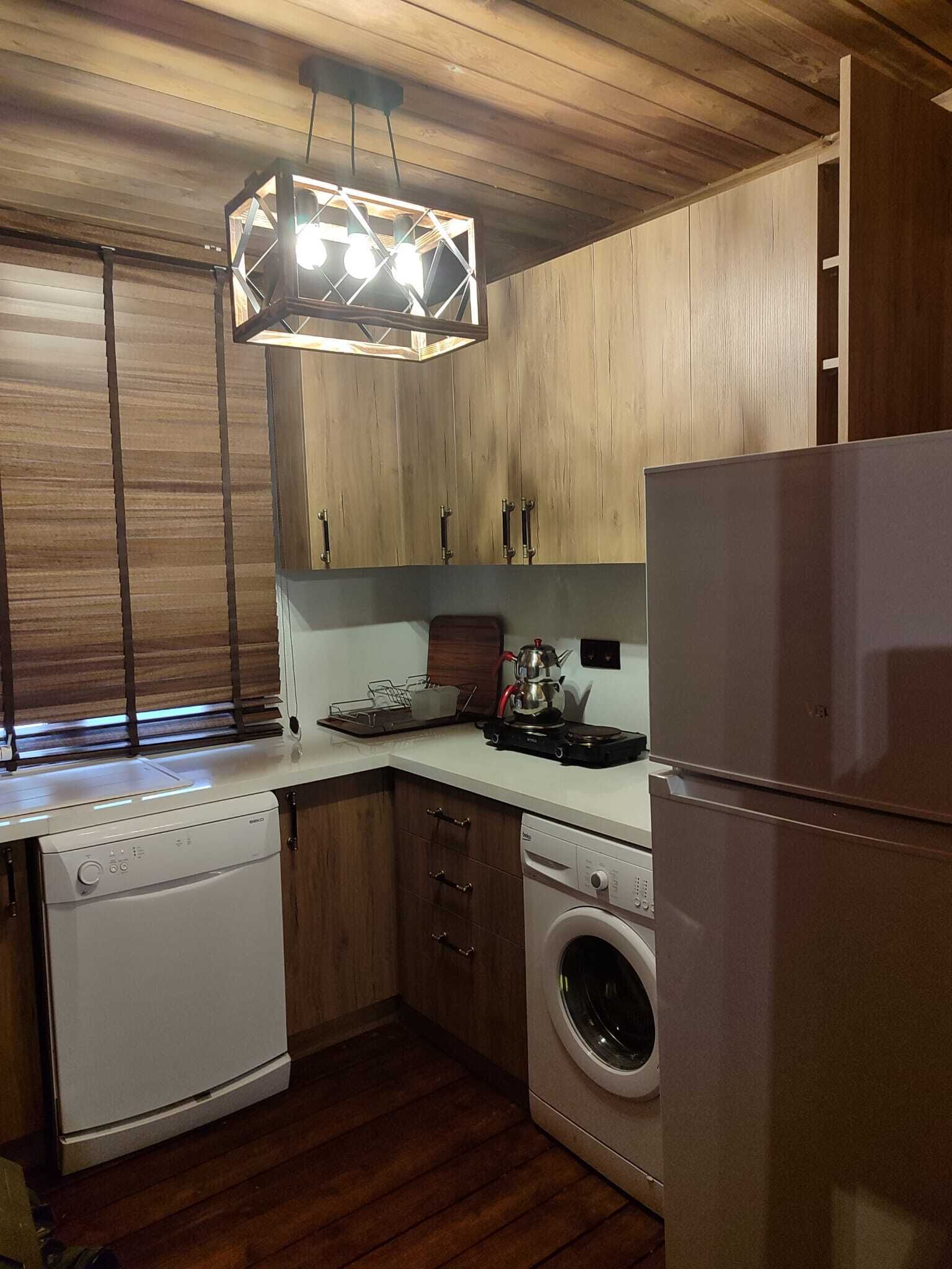 A compact kitchen with wooden cabinets, a built-in dishwasher, a washing machine, and a modern pendant light fixture.