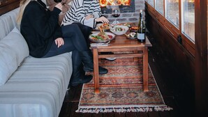 Interior acogedor de una cabaña con chimenea de ladrillo, paredes de madera, un sofá y una mesa de comedor con comida y bebidas.