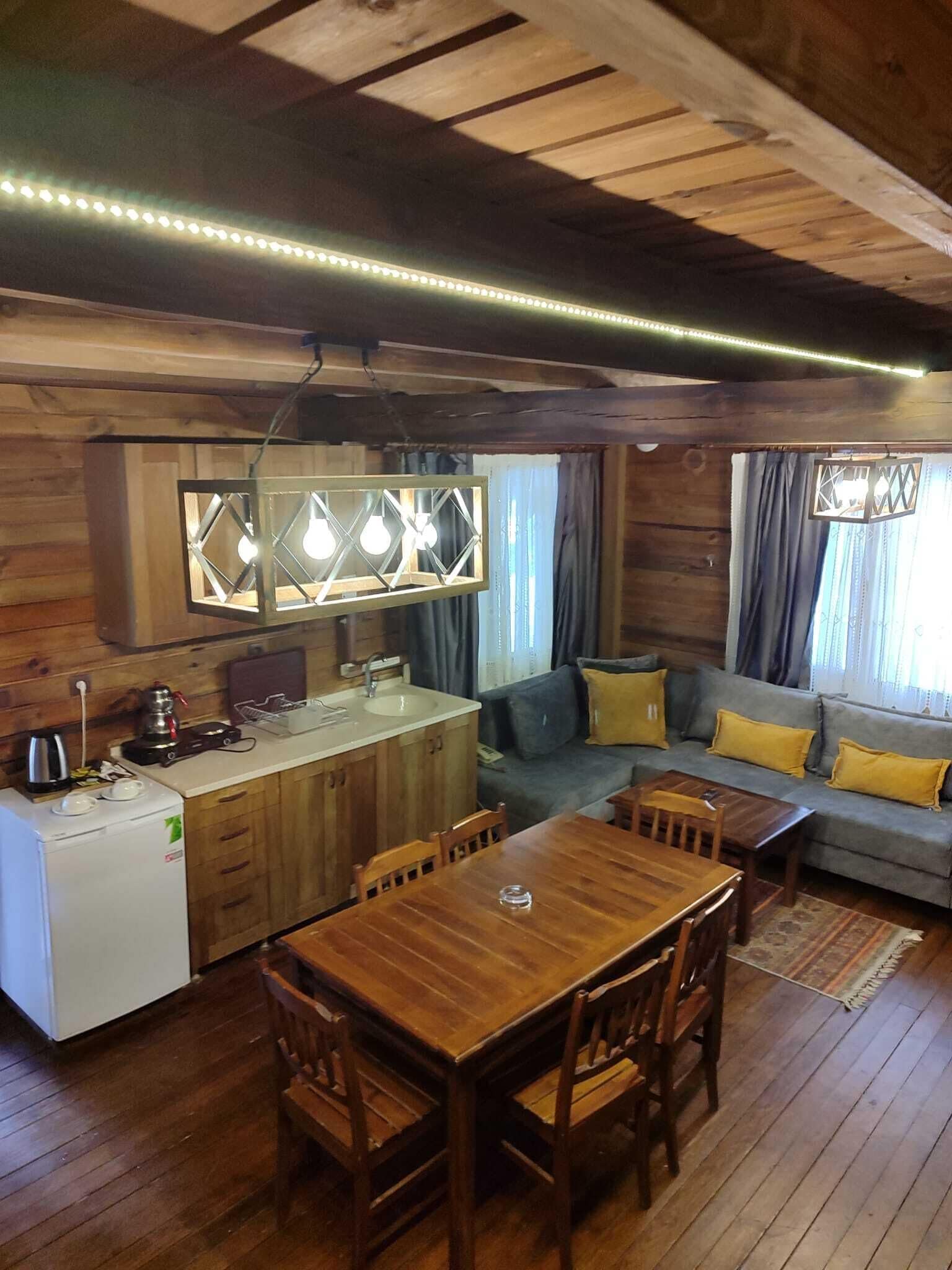Interior acogedor de una cabaña con una mesa de comedor de madera, sillas, una kitchenette y un sofá con cojines amarillos.