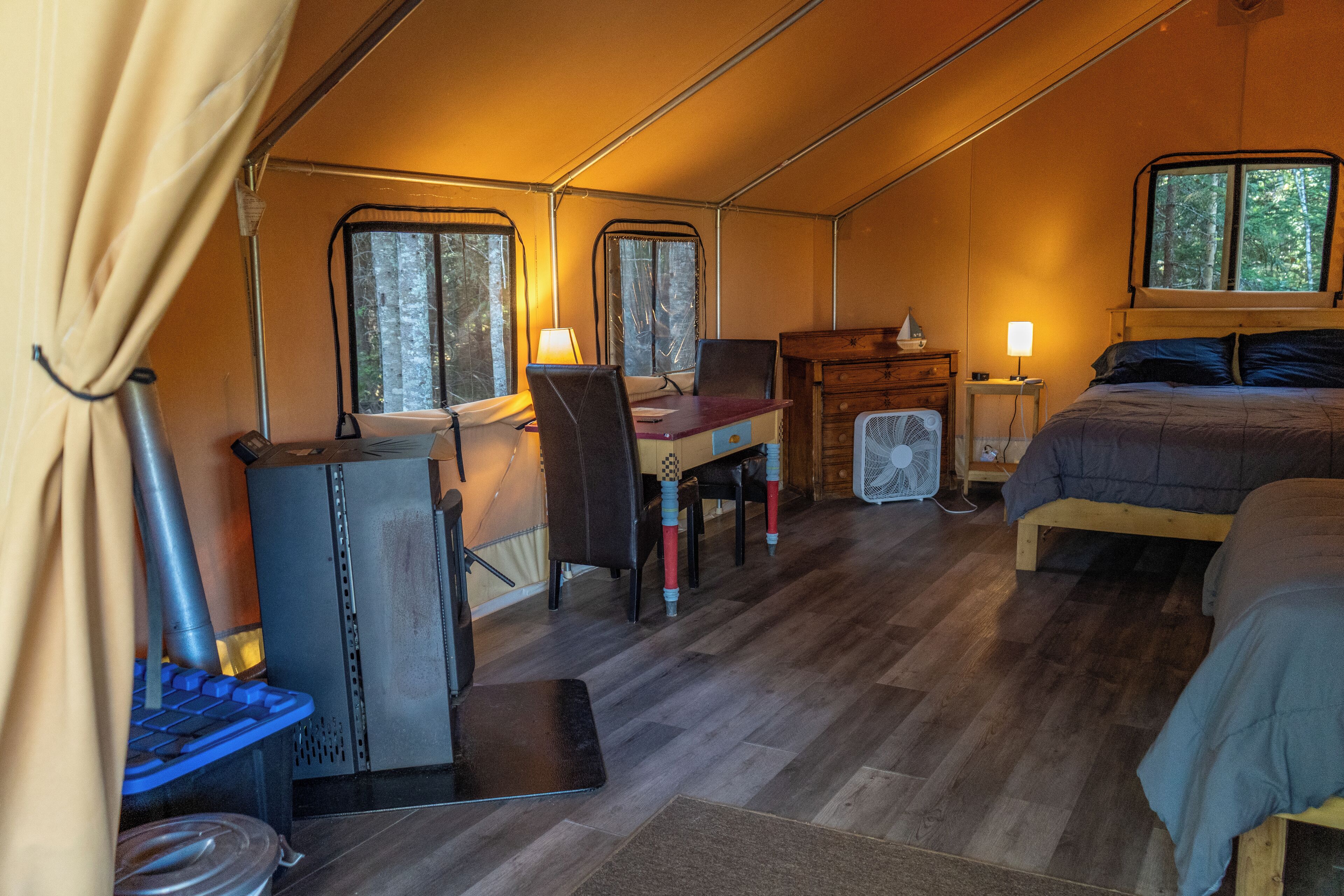A tent with a wooden floor, a desk with a chair, a bed, and a fan.