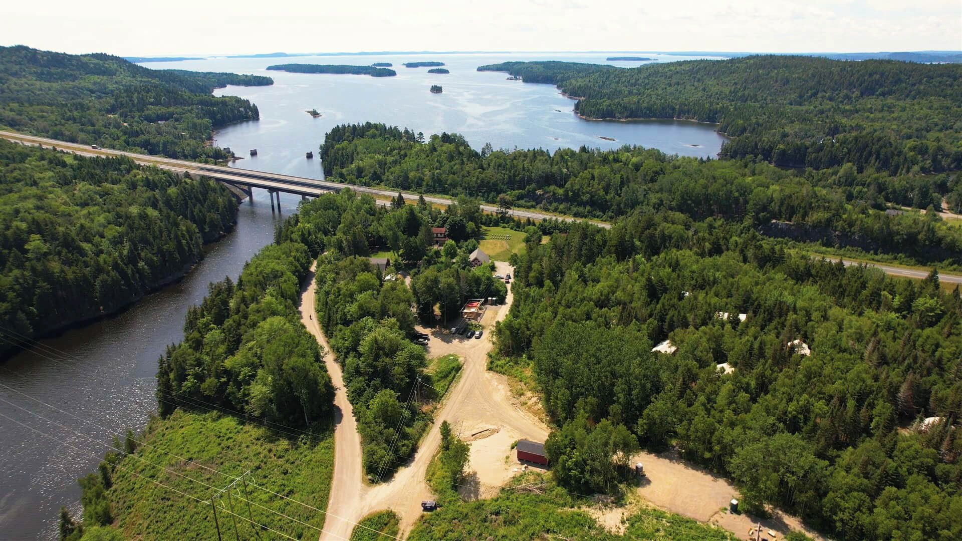 A riverside property with a dirt road, a bridge, and dense forest.