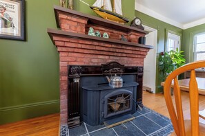 Una chimenea de ladrillo con un modelo de barco y un reloj encima, una silla de madera en primer plano y una pared verde con un cuadro enmarcado.