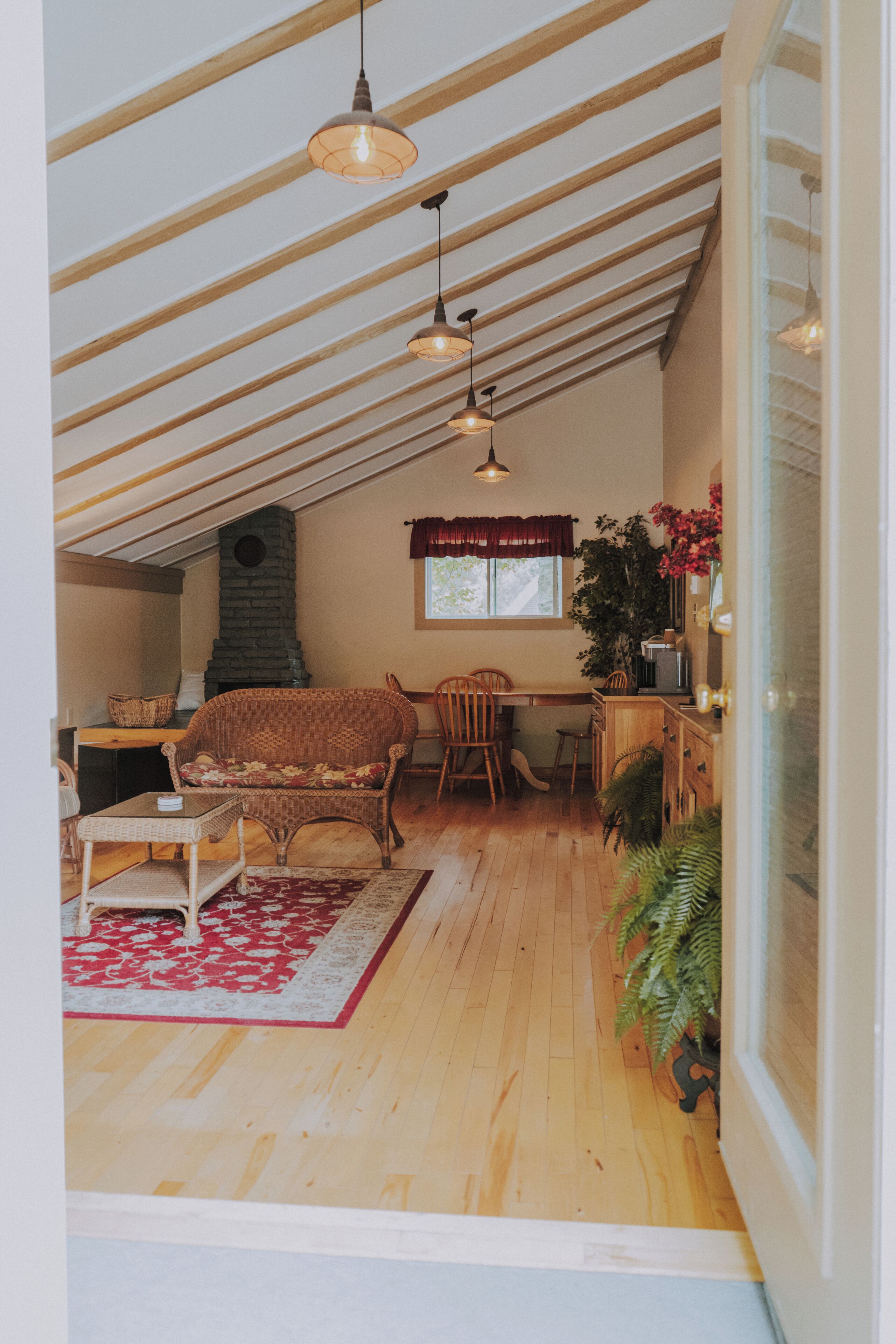 A cozy loft-style room with a sloped ceiling, wooden floors, a fireplace, wicker furniture, and hanging pendant lights.