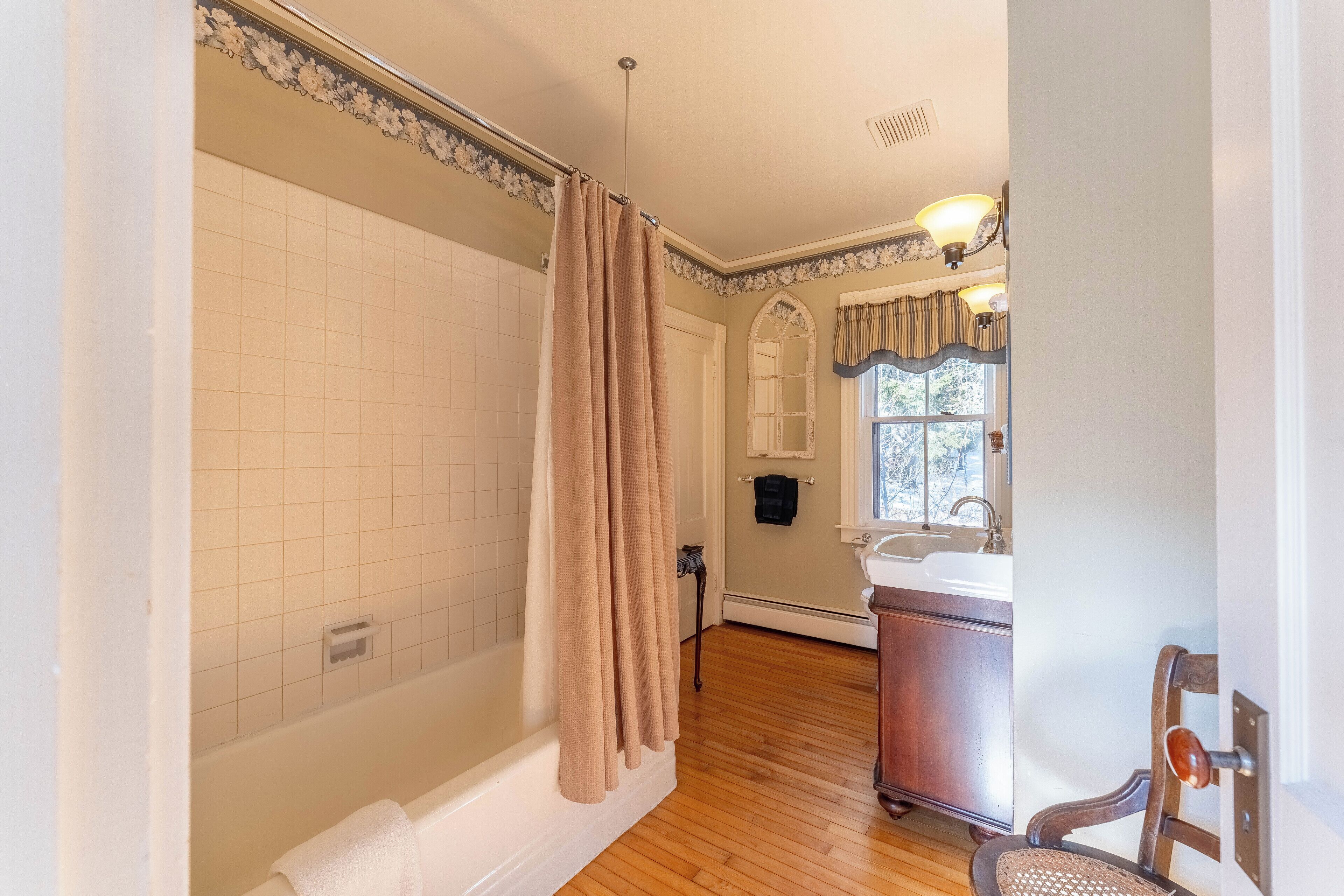 A bathroom with a bathtub, shower curtain, wooden floor, sink, and a chair.