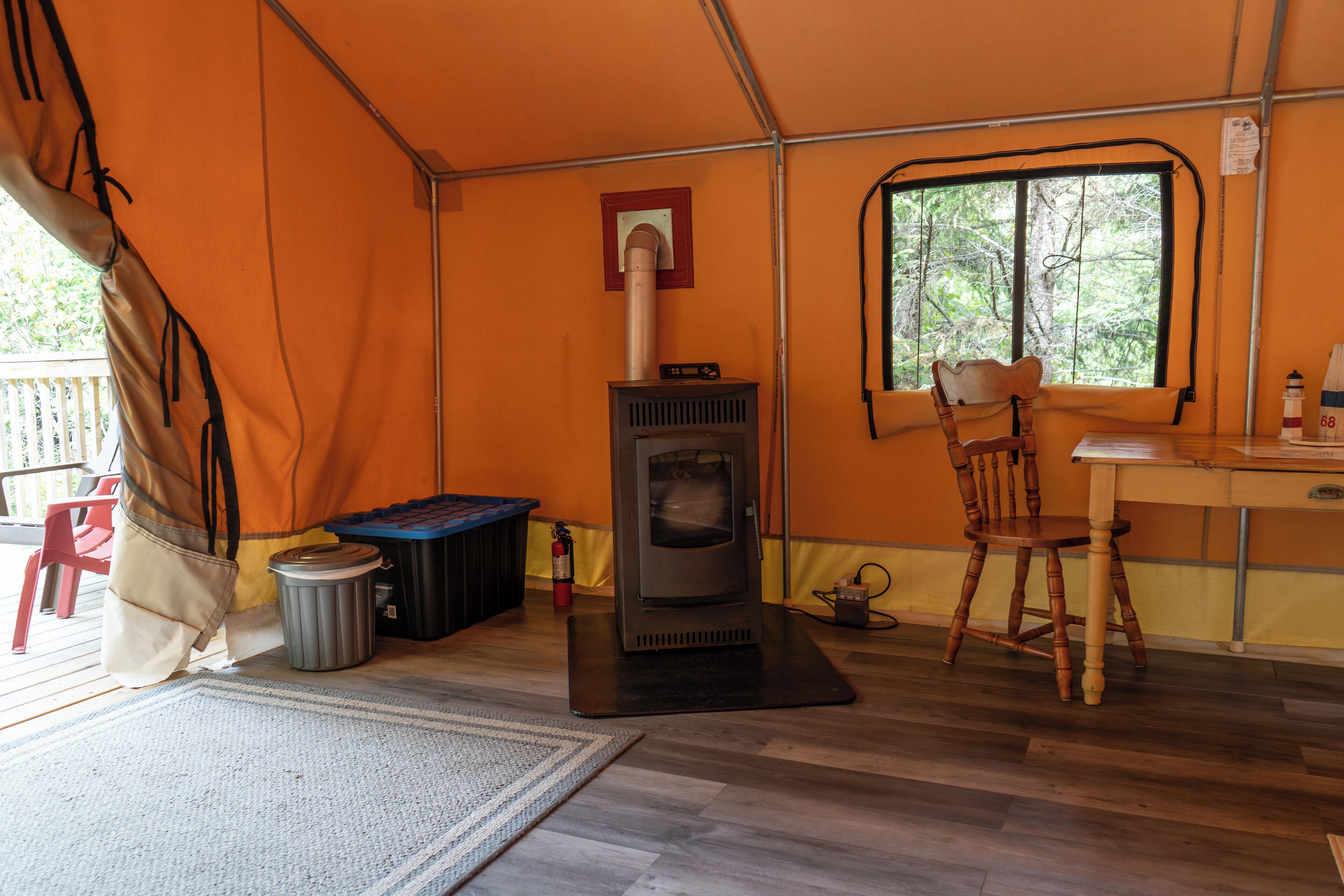 A tent with a wood stove, a chair, a table, and a window with a view of trees.