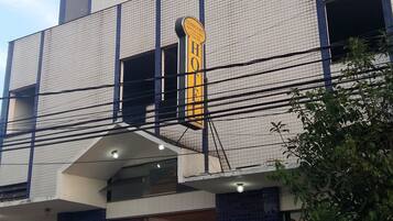 Um hotel com design moderno, apresentando uma placa amarela destacada com o nome 'COMFORT HOTEL'. O edifício tem vários andares com varandas, e há linhas de energia visíveis acima.