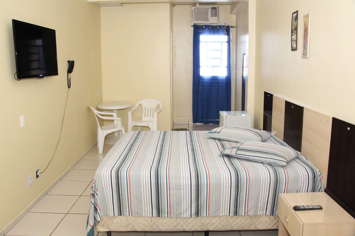 A hotel room with a bed, a TV mounted on the wall, a desk with a chair, and a window with a curtain.