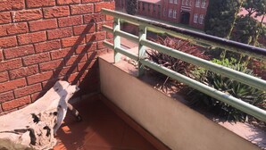 A balcony with a rustic wooden sculpture, a metal railing, and a view of a brick building and greenery. - Harare City 1-bed Apartment (Harare)