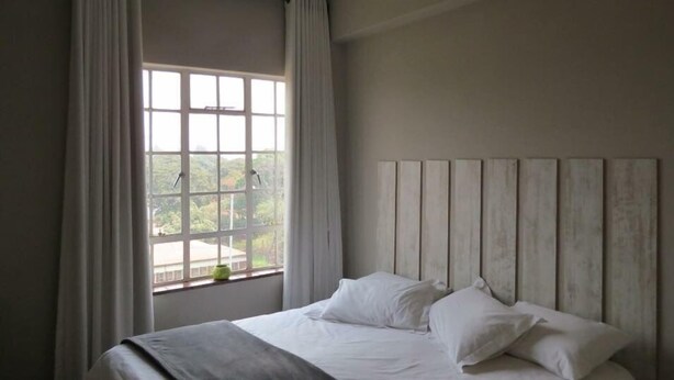A bedroom with a large window, a bed with white linens, and a wooden headboard. - Harare City 1-bed Apartment (Harare)