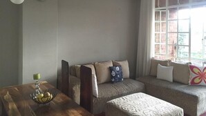 A cozy living room with a sofa, a wooden table with a fruit bowl, and a window with curtains. - Harare City 1-bed Apartment (Harare)