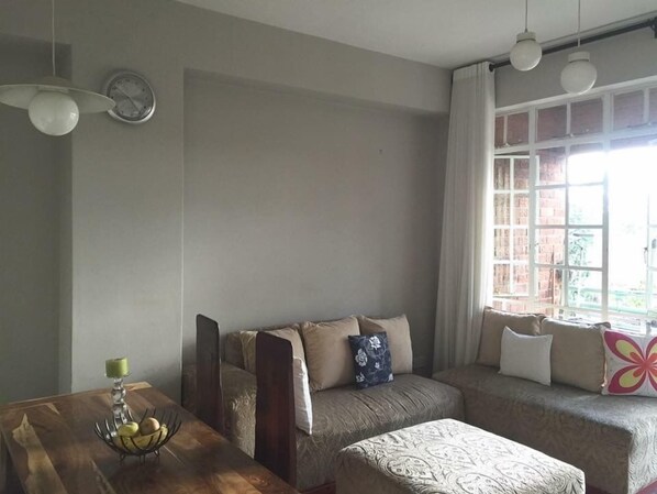 A cozy living room with a sofa, a wooden table with a fruit bowl, and a window with curtains.