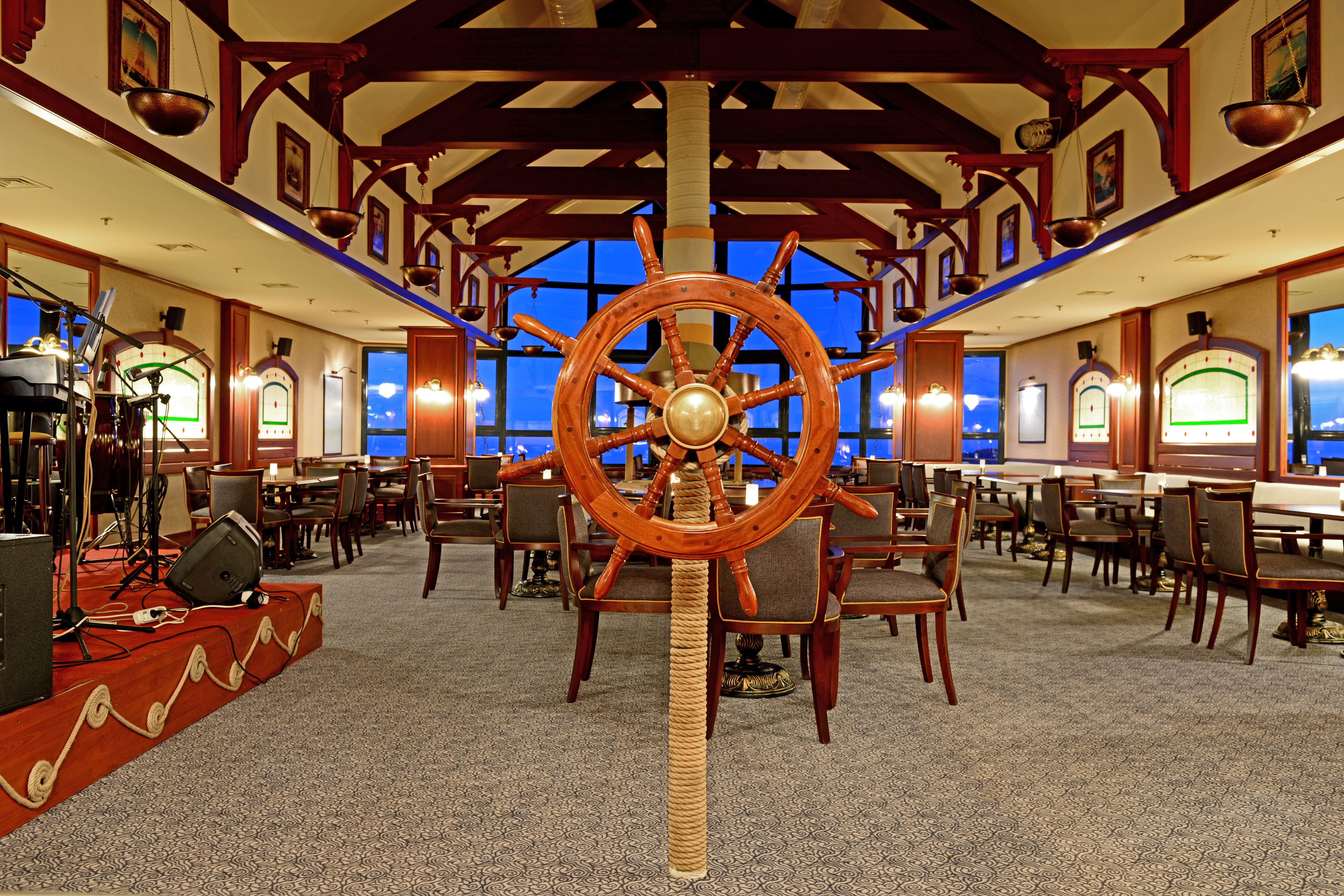 A large dining area with wooden tables and chairs, a prominent ship's wheel, and a stage with musical instruments.