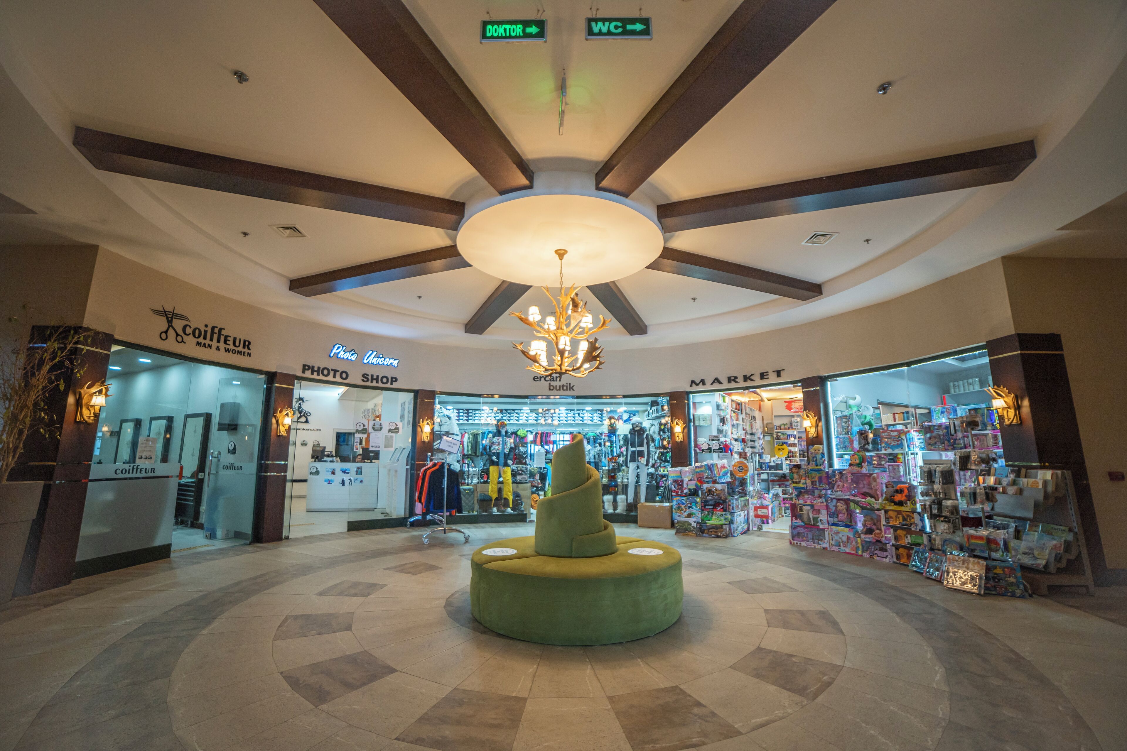 L’intérieur d’un centre commercial présente une sculpture centrale en forme de spirale verte, un plafond avec des poutres en bois et divers magasins, notamment un magasin de photographie et une épicerie.