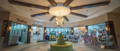 L’intérieur d’un centre commercial présente une sculpture centrale en forme de spirale verte, un plafond avec des poutres en bois et divers magasins, notamment un magasin de photographie et une épicerie.