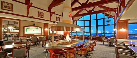 A spacious dining area with a central fire feature, wooden beams, large windows, and a view of the outside.