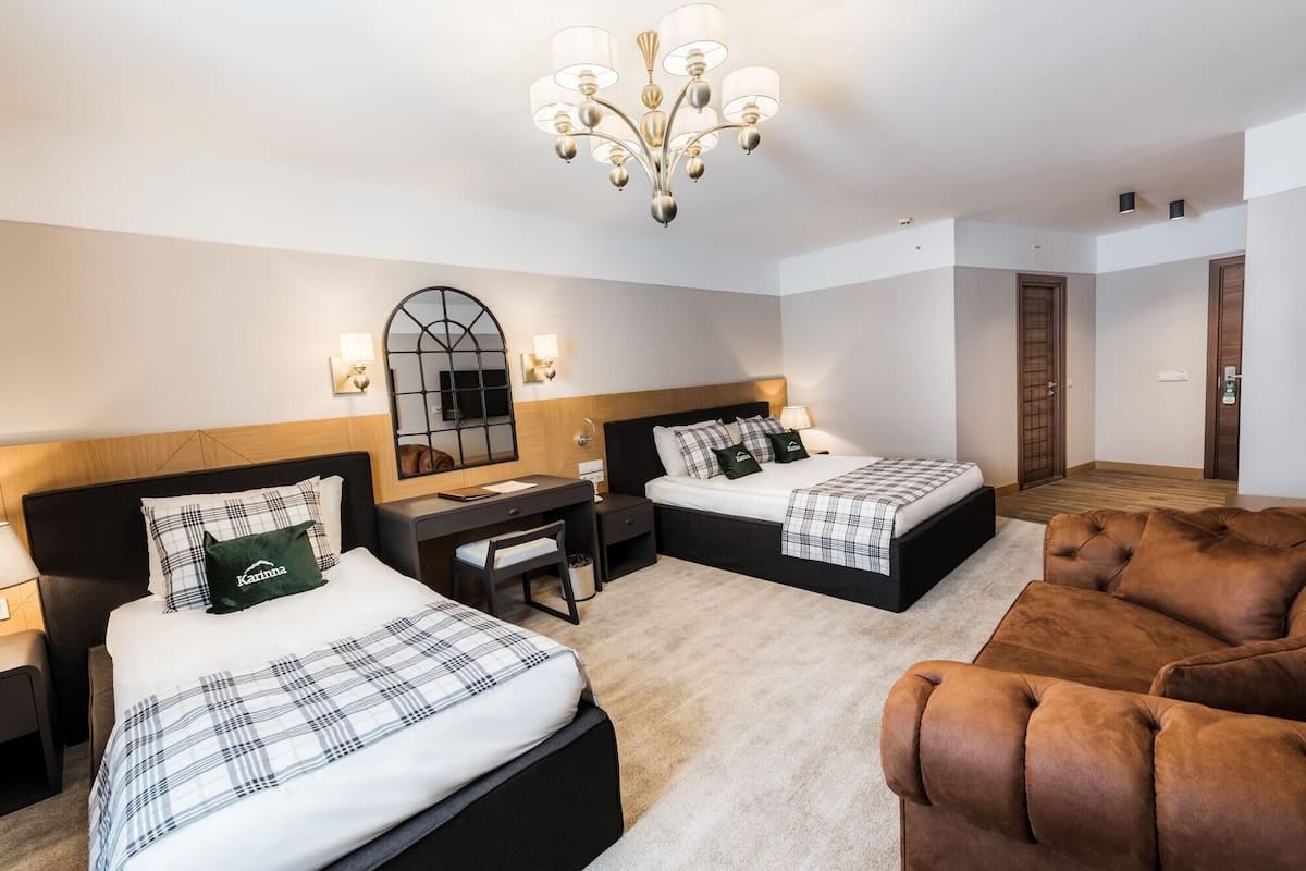 Une chambre d’hôtel avec deux lits, un bureau, un miroir, un lustre et un fauteuil marron.