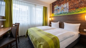 A hotel room with a bed, a desk, and a chair. There is a window with curtains, a painting on the wall, and a wooden headboard. - Trip Inn Hotel Münster City (Münster)